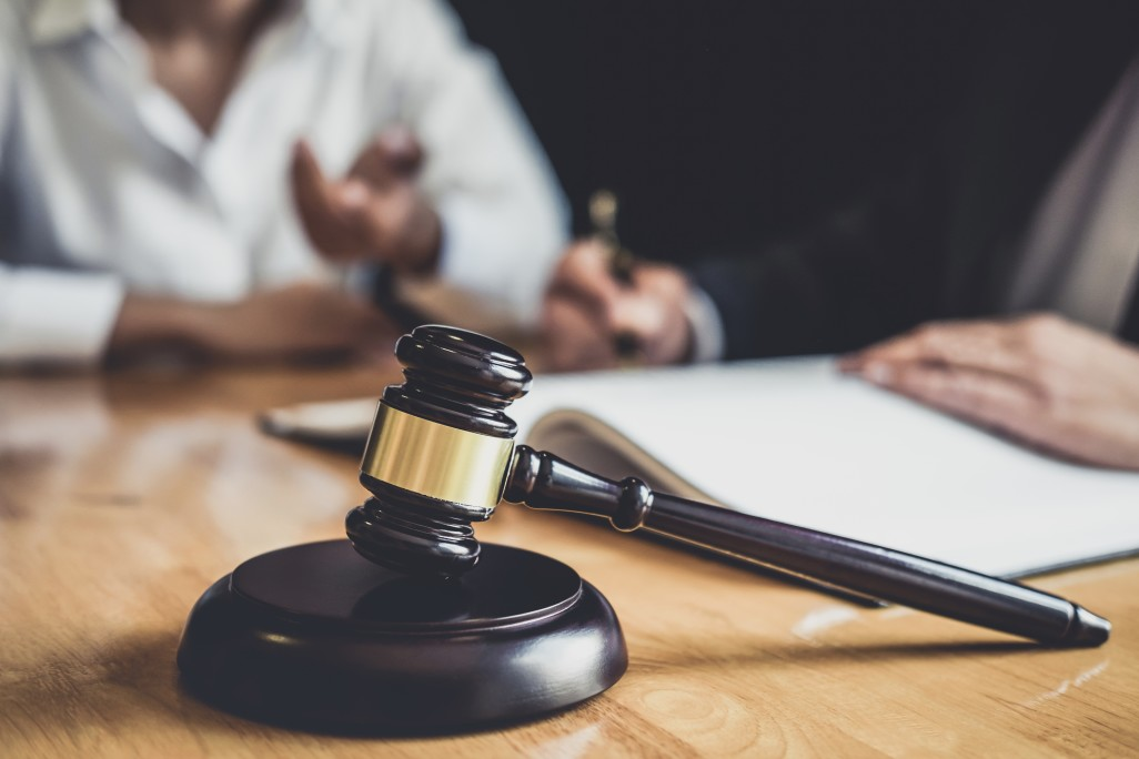 A gavel on a desk in the foreground, with two people having a legal discussion in the background.